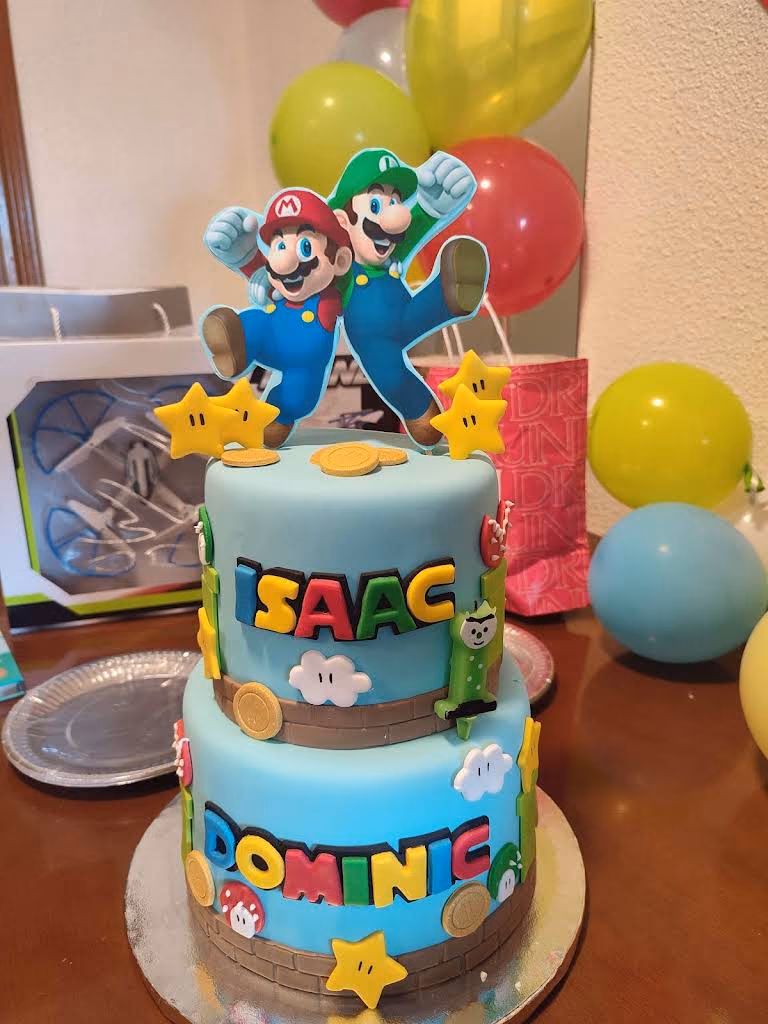 Tarta de dos pisos de cumpleaños con figuras y los nombres Isaac y Dominic rotulados en fondant